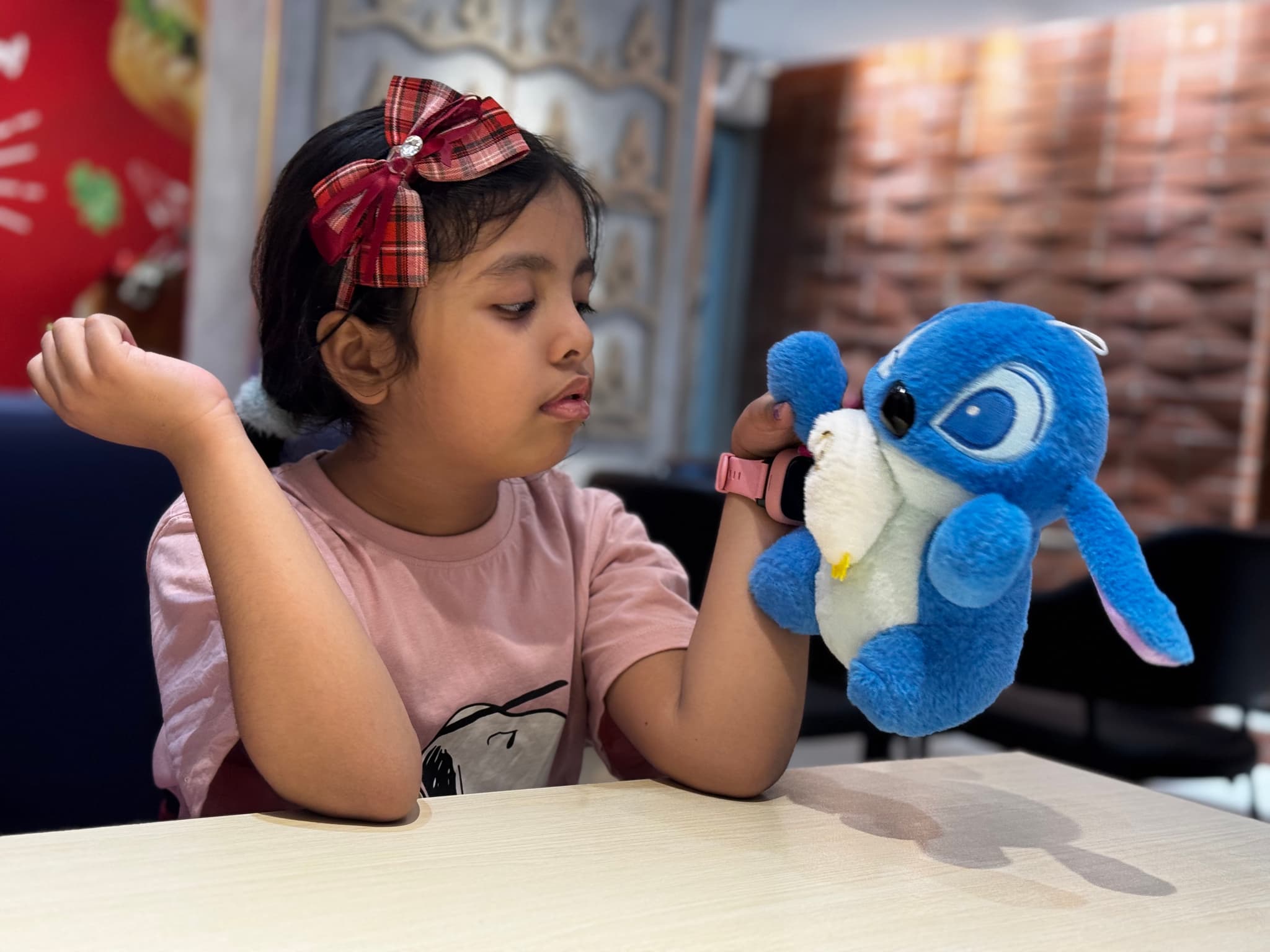 The image shows a young girl sitting at a table, holding a blue stuffed animal. She appears to be looking at the camera with a neutral expression. Behind her, there's a wall-mounted framed certificate and a decorative item that looks like a traditional Japanese lantern. The setting suggests it could be an interior space such as a home or a small office.