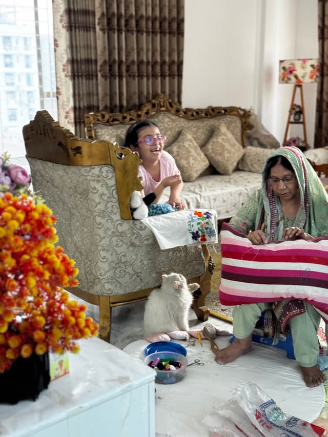 The image shows an indoor setting, with two individuals engaged in different activities. One person appears to be sorting through items on a surface, while another person is sitting on a couch and looking at something that is not visible. In the foreground, there are colorful flowers placed on surfaces. There is also a pet cat resting nearby. The room has various decorative elements such as potted plants, vases with flowers, and wall hangings. The walls are decorated in a traditional manner, suggesting an Indian or South Asian context. Natural light seems to be entering the room from a window out of frame, illuminating the scene.