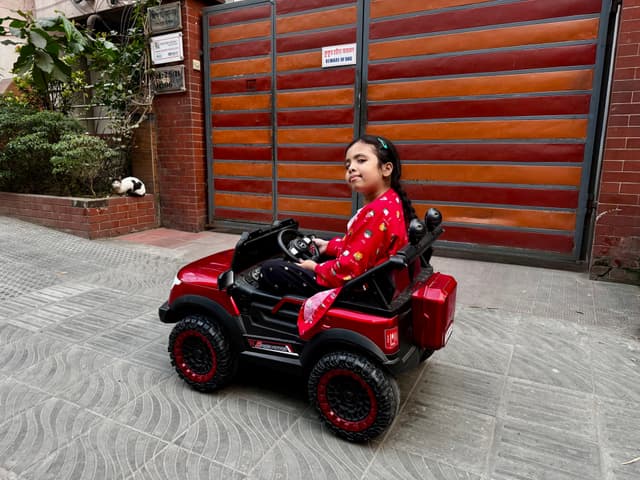The image displays a child sitting in a red four-wheeler outdoors. They appear to be wearing a dark long-sleeved shirt and are positioned towards the front of the vehicle, looking directly at the camera with a neutral expression. Behind the child, there's a closed garage door, suggesting that they might be near a residential area. The photo has two versions in side by side: one where the image is normal and another where it's mirrored, possibly to demonstrate how the same scene might look to someone from a region where vehicles drive on the left or right side of the road.
