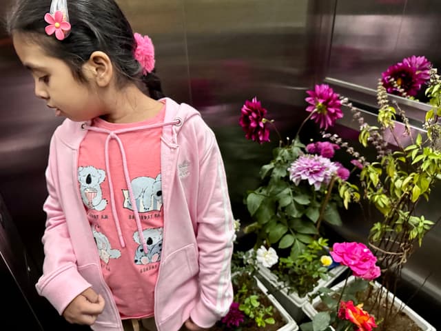 The image features a young girl with a flower arrangement in the foreground. She is standing and looking down at the flowers. Behind her, there are plants and flowers visible through a glass door. The background appears to be an indoor space, possibly a balcony or garden area, with potted plants and flowers arranged for display. The lighting suggests it could be daytime due to the natural light illuminating the scene.